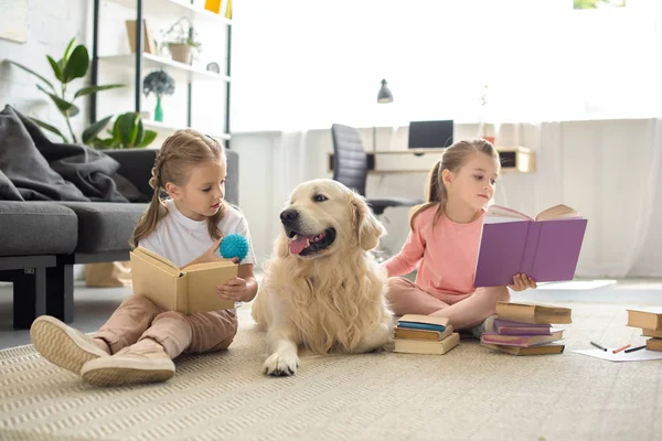 Hermanas pequeñas leyendo libros con perro golden retriever cerca en casa - foto de stock