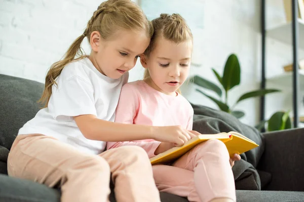 Adorables hermanitas leyendo el libro juntas en el sofá en casa - foto de stock