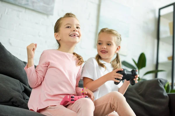 Retrato de hermanas pequeñas con gamepads jugando videojuego juntas en casa - foto de stock