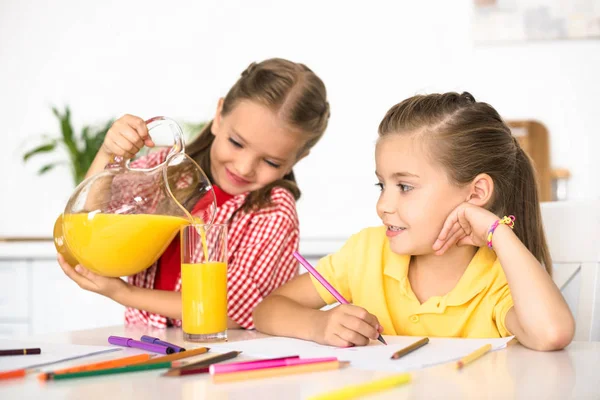 Retrato de niño vertiendo jugo en el vaso para hermana en la mesa con papeles y lápices para dibujar en casa - foto de stock