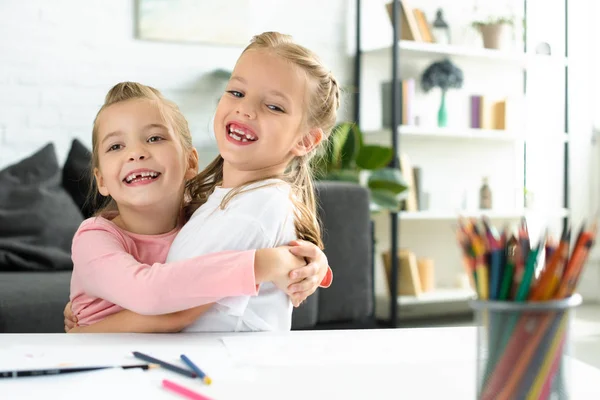 Portrait de petits enfants heureux s'embrassant à table avec des papiers et des crayons pour dessiner à la maison — Photo de stock