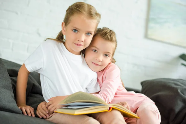 Ritratto di bambina abbracciata sorella con libro sul divano di casa — Foto stock