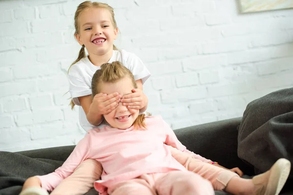 Entzückendes kleines Kind, das seiner Schwester auf dem heimischen Sofa die Augen verdeckt — Stockfoto