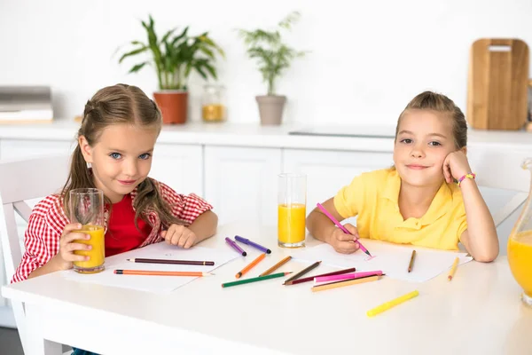 Retrato de lindos niños sonrientes mirando a la cámara mientras dibuja imágenes en la mesa en casa - foto de stock