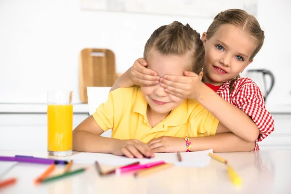 Mignon petit enfant couvrant les yeux de sœur à table avec des crayons et des papiers pour le dessin à la maison — Photo de stock