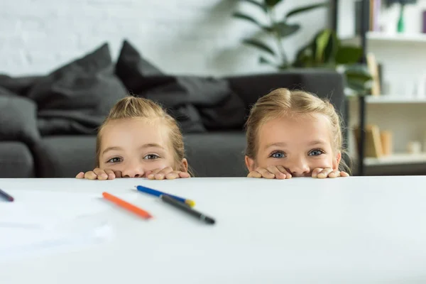 Vista parcial de los niños pequeños mirando a la cámara mientras se esconden detrás de la mesa con lápices en casa - foto de stock