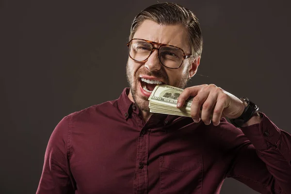 Empresario emocional en gafas mordiendo billetes de dólar aislados en negro - foto de stock