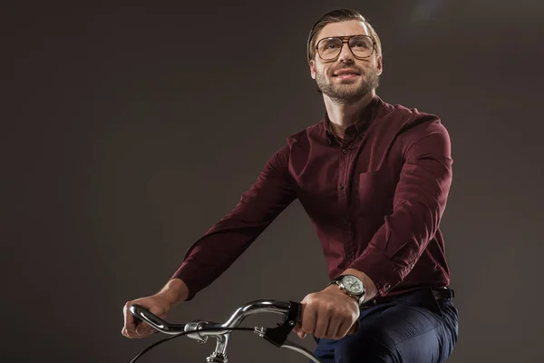 Low angle view of smiling man in eyeglasses riding bike and looking away isolated on black — Stock Photo