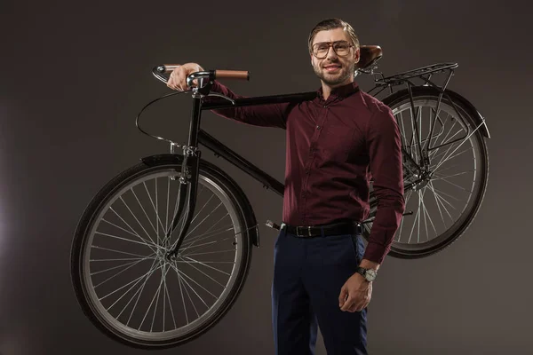 Handsome man in eyeglasses holding bicycle and smiling at camera on black — Stock Photo