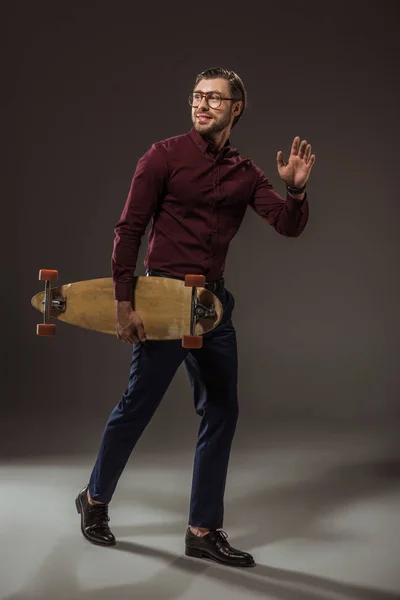 Ganzkörperansicht eines gut aussehenden lächelnden Mannes mit Brille, der Longboard hält und mit der Hand auf schwarz wedelt — Stockfoto