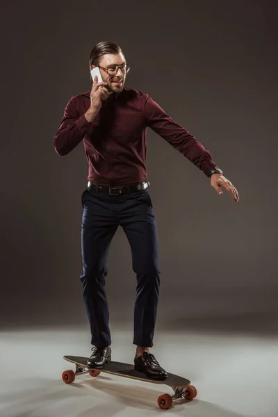 Улыбающийся бизнесмен разговаривает по смартфону, стоя на скейтборде и глядя в сторону черного — стоковое фото