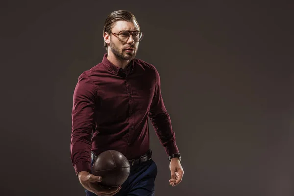 Красивый мужчина в очках, держит регби-мяч и смотрит в сторону, изолированный от черного — стоковое фото