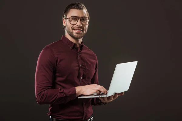 Bell'uomo con gli occhiali usando il computer portatile e sorridendo alla fotocamera isolata sul nero — Foto stock