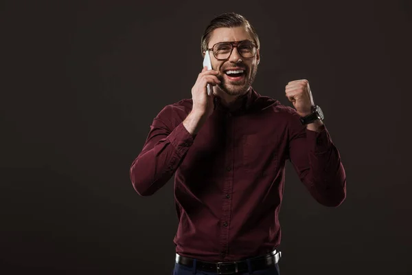 Fröhlicher Mann mit Brille, der mit dem Smartphone spricht und isoliert auf schwarz triumphiert — Stockfoto