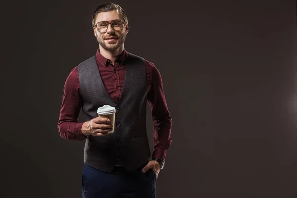 Красивий чоловік в окулярах тримає одноразову чашку кави і посміхається на камеру ізольовано на чорному — стокове фото