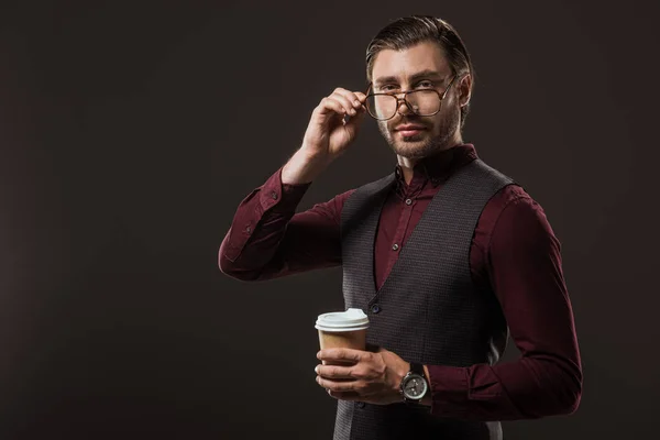 Elegante hombre de negocios con café para ir ajustando las gafas y mirando a la cámara aislada en negro - foto de stock