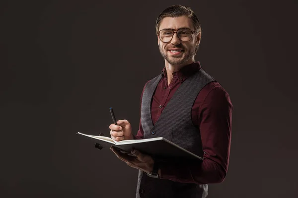 Handsome buisnessman in eyeglasses taking notes and smiling at camera isolated on black — Stock Photo