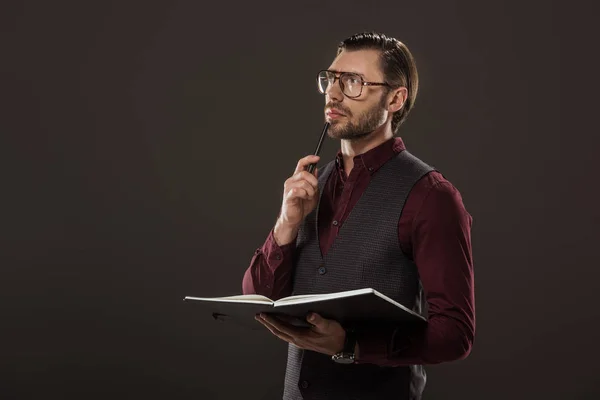 Empresário pensativo em óculos segurando notebook e caneta isolada em preto — Fotografia de Stock