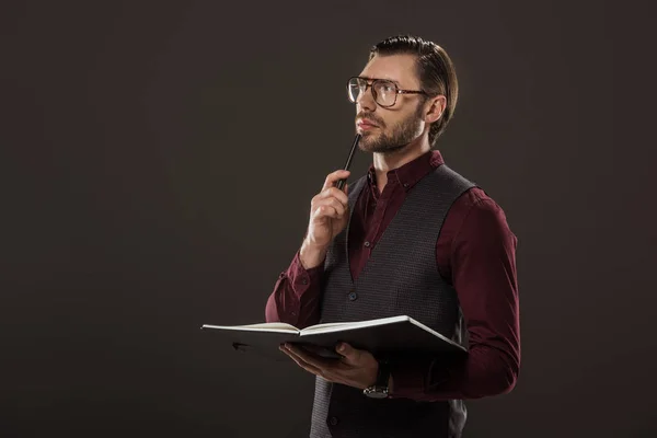 Pensive businessman in eyeglasses holding notebook and pen isolated on black — Stock Photo