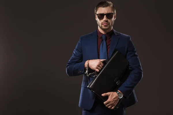 Businessman in sunglasses and handcuffs holding briefcase and looking at camera isolated on black — Stock Photo