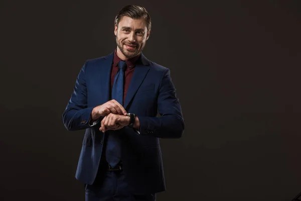 Handsome businessman checking wristwatch and smiling at camera isolated on black — Stock Photo