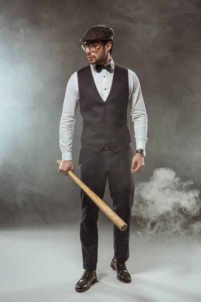 Вид в полный рост стильного мужчины в галстуке, очках и кепке, держащего бейсбольную биту и отводящего взгляд в дыму — стоковое фото