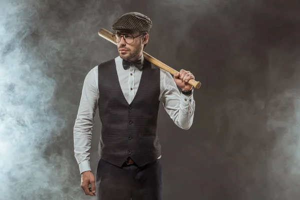 Hombre con estilo en corbata de lazo, gafas y gorra sosteniendo bate de béisbol y mirando hacia otro lado en negro — Stock Photo