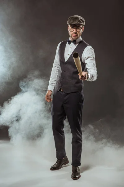 Ernster, stylischer Mann mit Fliege und Mütze hält Baseballschläger in der Hand, während er im Rauch steht — Stockfoto