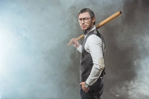 Homem elegante sério segurando bastão de beisebol e olhando para a câmera enquanto em pé na fumaça — Fotografia de Stock