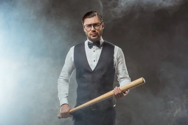 Hombre guapo con pajarita y anteojos sosteniendo bate de béisbol y mirando a la cámara en humo - foto de stock
