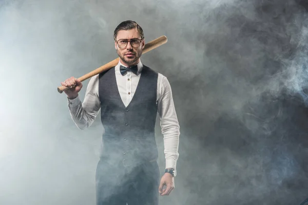 Hombre con estilo en pajarita y gafas con bate de béisbol y mirando a la cámara en el humo - foto de stock