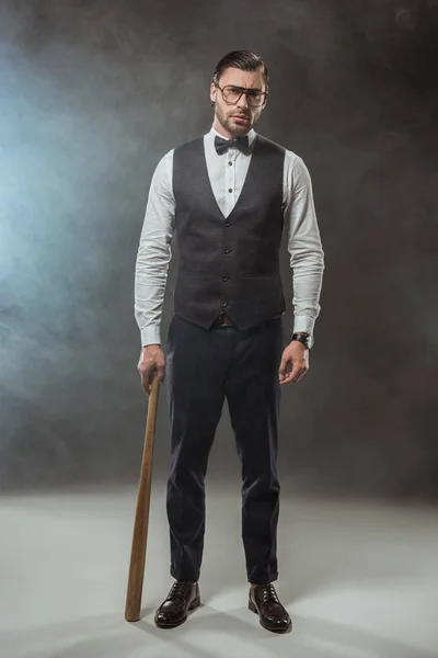 Vista completa de hombre con estilo en pajarita y gafas con bate de béisbol y mirando a la cámara en el humo - foto de stock