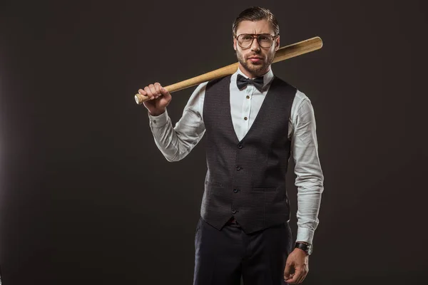 Stylish man in bow tie and eyeglasses holding baseball bat and looking at camera isolated on black — Stock Photo