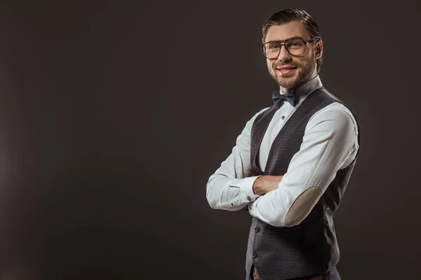 Portrait of handsome stylish man in bow tie and eyeglasses standing with crossed arms and smiling at camera isolated on black — Stock Photo