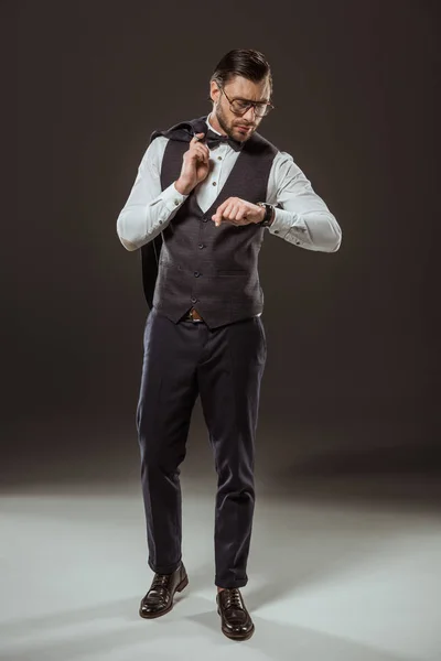 Vue pleine longueur de l'homme d'affaires élégant en noeud papillon et lunettes tenant veste costume et vérifier la montre-bracelet sur noir — Photo de stock