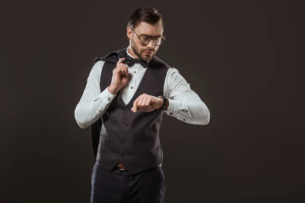 Стильный бизнесмен в галстуке-бабочке и очках, держащий пиджак и проверяющий наручные часы, изолированные на черном — стоковое фото