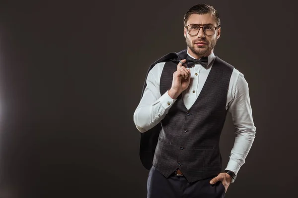 Handsome stylish man in bow tie and eyeglasses holding suit jacket and looking at camera isolated on black — Stock Photo
