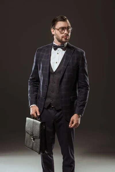 Buisnessman guapo en traje, anteojos y pajarita sosteniendo maletín y mirando hacia otro lado en negro - foto de stock