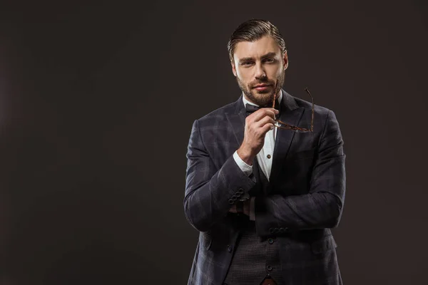 Handsome man in suit and bow tie holding eyeglasses and looking at camera isolated on black — Stock Photo