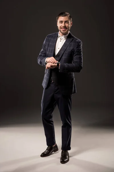Vista de longitud completa de hombre de negocios con estilo comprobar reloj de pulsera y sonriendo a la cámara en negro - foto de stock