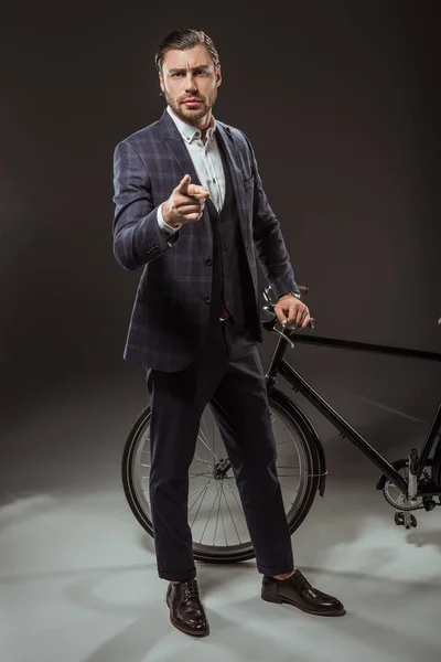 Serious young man in suit pointing at camera while standing near bicycle on black — Stock Photo