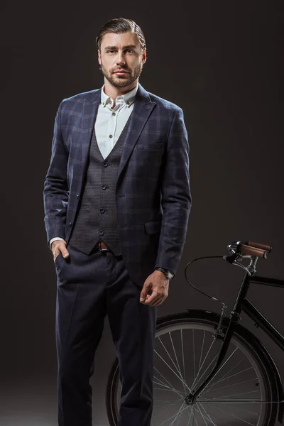 Красивий молодий чоловік у костюмі дивиться на камеру, стоячи біля велосипеда на чорному — стокове фото