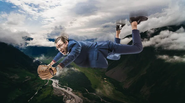 Homem de negócios em óculos com luva de beisebol voando em nuvens acima da paisagem natural incrível — Fotografia de Stock