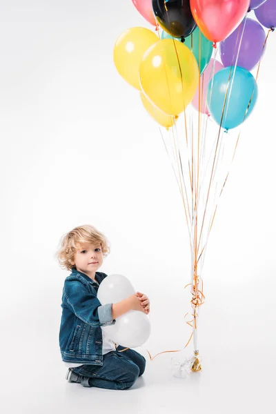 Adorabile ragazzo seduto con fascio di palloncini su bianco — Foto stock