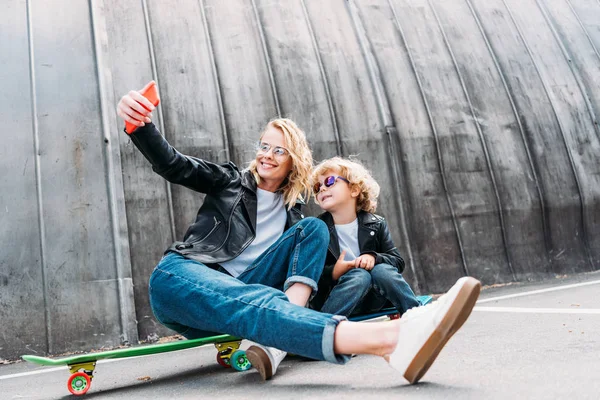 Madre e hijo sentados en monopatines en la calle y tomando selfie con smartphone - foto de stock