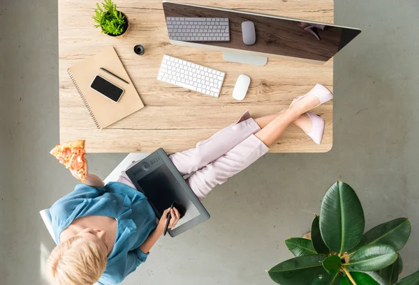 Top view of stylish female freelancer eating pizza and using graphic tablet with legs on table at home — Stock Photo