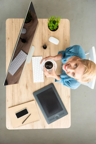 Vista elevata di una donna freelance sorridente che guarda la macchina fotografica e tiene in mano una tazza di caffè a tavola con tablet grafico, smartphone e computer a casa ufficio — Foto stock