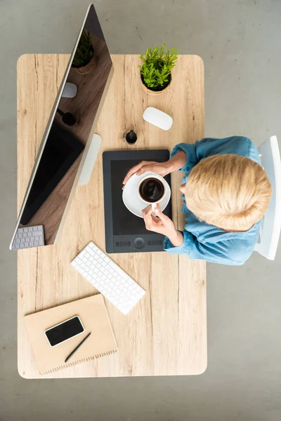 Vue aérienne d'une pigiste tenant une tasse de café à table avec tablette graphique, smartphone et ordinateur au bureau à domicile — Photo de stock