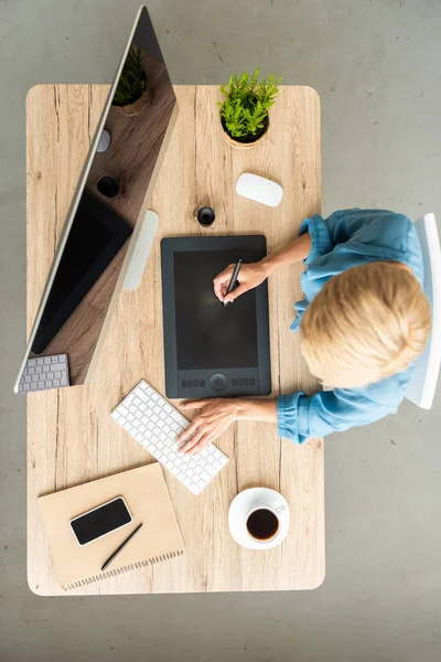 Вид сверху женщины-фрилансера, работающей на графическом планшете за столом с чашкой кофе, смартфоном и компьютером в домашнем офисе — стоковое фото