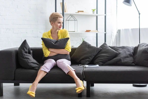 Lächelnde junge Frau mit Kopfkissen in der Hand und zu Hause auf dem Sofa sitzend mit digitalem Tablet und Smartphone — Stockfoto